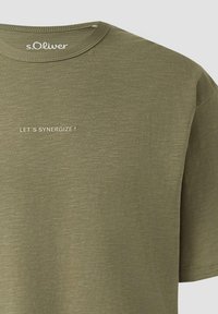T-shirt olive s.Oliver avec col rond et texte « LET'S SYNERGIZE ! » imprimé sur le côté gauche de la poitrine.