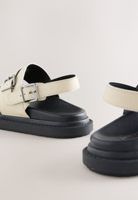 Sandales compensées beige et noir avec brides ajustables à boucle et semelles épaisses noires sur fond neutre.