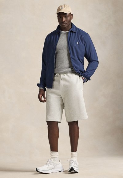 Navy lightweight jacket, gray sweater, light gray shorts, white sneakers with pattern, beige cap. Model poses with hands in pockets.
