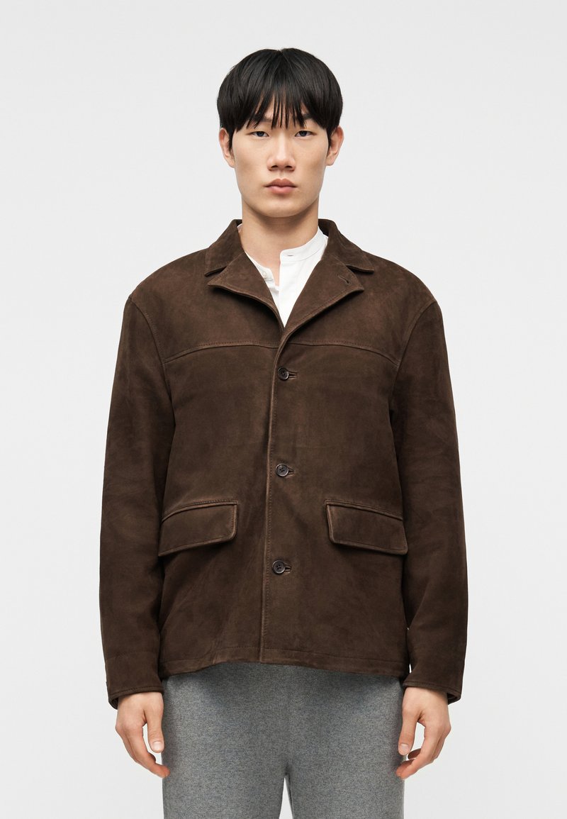 Νεαρός άνδρας που φοράει σκούρο κα φέ σουέντ μπουφάν πάνω από λευκό πουκάμισο και γκρι παντελόνι, στέκεται μπροστά σε απλό φωτεινό φόντο.