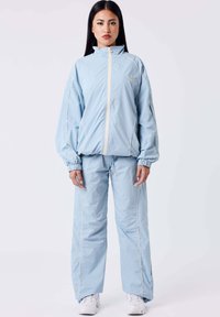 Tuta azzurro chiaro realizzato in tessuto liscio e leggero, caratterizzato da un colletto alto, chiusura a zip e polsini elasticizzati, con dettagli in piping bianco a contrasto.