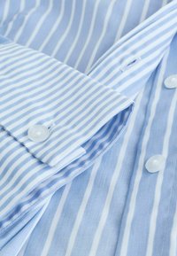 Chemise en coton bleu clair avec des rayures blanches, dotée d'une fermeture à boutons et d'une texture lisse, présentant une manche repliée pour le détail.
