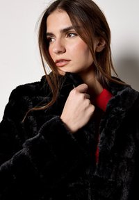 Czarna kurtka z sztucznego futra z wysokim kołnierzem, o puszystej fakturze i szerokich rękawach. Pod spodem częściowo widoczny jest czerwony sweter dziany.