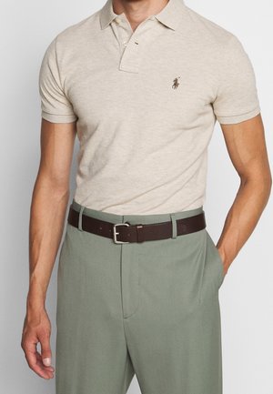 Beige polo met korte mouwen en geborduurd logo, gecombineerd met lichtgroene broek en een donkerbruine leren riem.