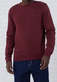 Felpa bordeaux con collo a giro e maniche lunghe, caratterizzata da una finitura testurizzata e un piccolo logo nero sul petto.