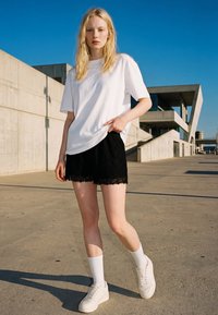 Blond naine valges t-särgis, mustades lühikestes pükstes, valgete sokkide ja tossudega seisab päikeselisel päeval tänapäevase betoonhoone juures õues.