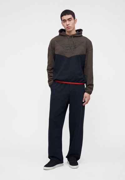 Jeune homme debout portant un sweat à capuche bicolore marron et noir avec logo, pantalon large noir, baskets noires et une chemise rouge dépassant.