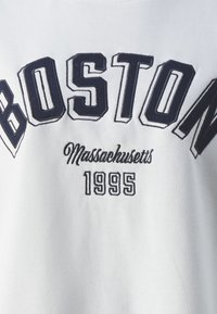 Sudadera blanca con texto bordado en azul que dice "BOSTON" en una fuente cuadrada, "Massachusetts" en cursiva, y "1995" debajo.