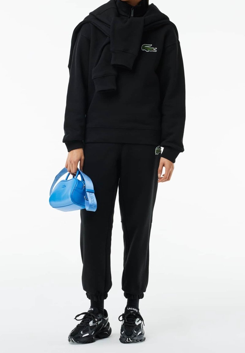 Zwarte hoodie en joggingbroek met een groen logo, gecombineerd met zwarte sneakers. Houdt een glanzende blauwe handtas met een blauwe band vast.