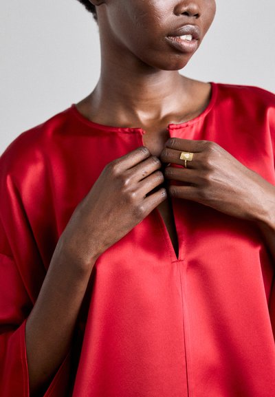 Blusa de satén roja con escote en V y mangas sueltas, que muestra una textura suave. Un anillo dorado adorna una mano que ajusta el cuello.