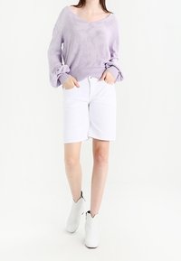 Lila pletený svetr s výstřihem do V a nafouklými rukávy, doplněný bílými šortkami ke kolenům a bílými kotníkovými botami s černým detailem.