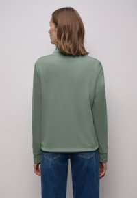Felpa verde con collo alto, maniche lunghe e texture liscia. Abbinata a jeans blu, che mostrano una vestibilità comoda e semplicità.
