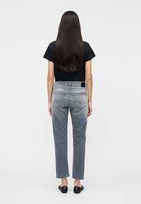 Femme aux longs cheveux foncés portant un t-shirt noir, un jean gris délavé avec coutures sur la poche arrière, et des chaussures noires sans lacets, debout de dos.
