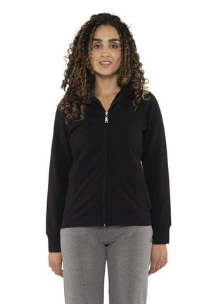 Une femme aux cheveux bouclés, vêtue d’un sweat à capuche noir zippé et d’un pantalon de jogging gris, se tient devant un fond blanc.