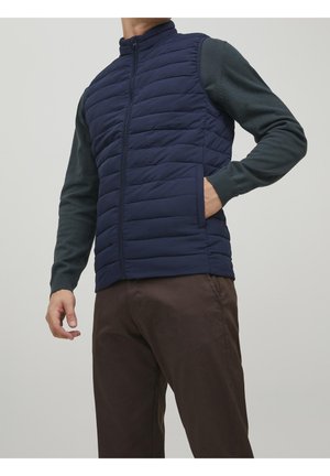 Man draagt een marineblauw doorgestikt vest met rits over een donkergroene trui en bruine broek, staand met één hand in het zakje van het vest.