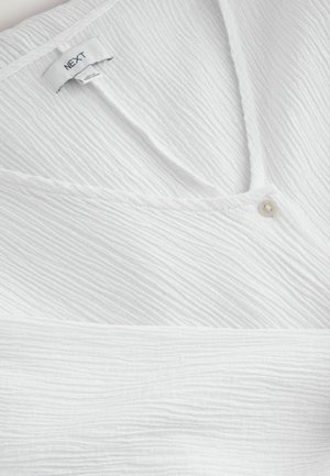 White textured blouse with a V-neck, featuring a small button accent. Fabric shows a wavy, crinkled pattern. Branded label visible inside.