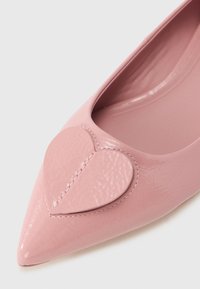 Ροζ παπούτσι μπαλαρίνα από δερματίνη με μυτερή μύτη. Διαθέτει καρδιά από δέρμα στο μπροστινό μέρος και λείο, γυαλιστερό φινίρισμα.