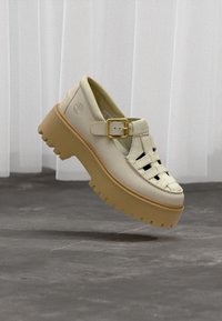 Δερμάτινο παπούτσι Mary Jane σε κρεμ απόχρωση με σόλα τύπου lug και χρυσό λουράκι με αγκράφα, αιωρούμενο πάνω από γκρι σκυρόδεμα και φόντο λευκής κουρτίνας.