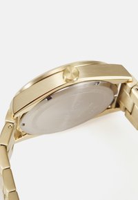 Reloj de metal en tono dorado con una superficie lisa y pulida. Presenta una caja redonda, una corona texturizada y un diseño de pulsera de eslabones metálicos.