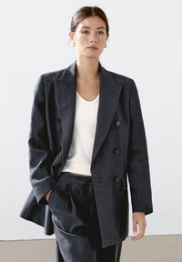 Blazer doppiopetto a righe navy, tessuto leggero, ampi revers, bottoni neri, abbinato a una maglietta bianca aderente e pantaloni coordinati.