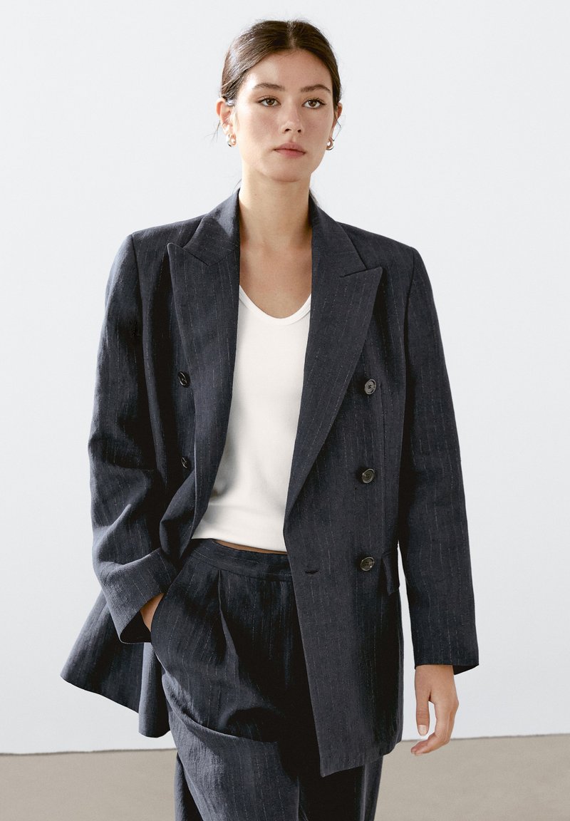 Blazer doppiopetto a righe navy, tessuto leggero, ampi revers, bottoni neri, abbinato a una maglietta bianca aderente e pantaloni coordinati.