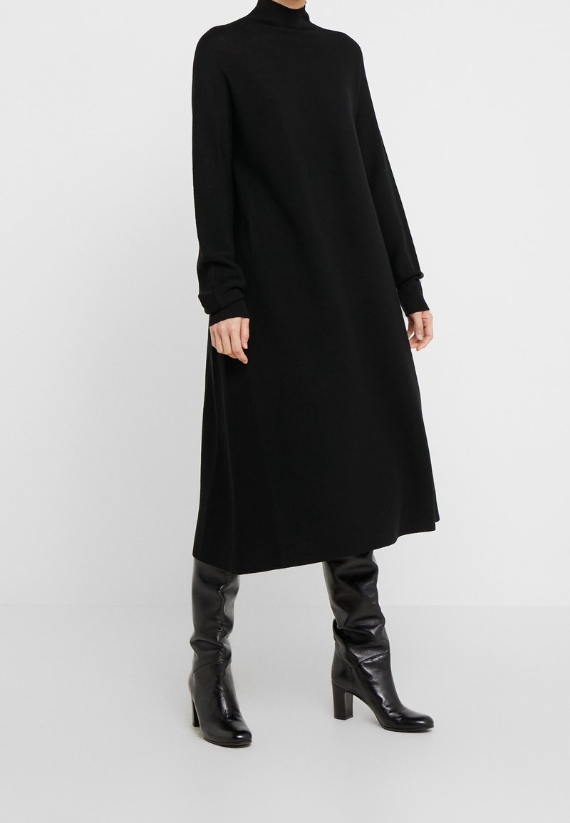 Femme portant une robe midi noire à manches longues et col roulé, associée à des bottes en cuir noires montant jusqu'aux genoux avec talons, sur fond blanc.