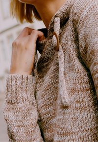 Femme ajustant le col d’un pull en maille beige et marron avec un gros bouton en bois près du cou.