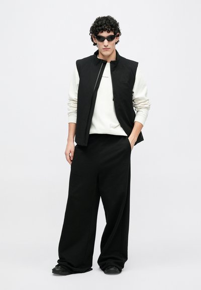 Fekete ujjatlan mellény fehér pulóver fölött, széles fekete nadrággal és fekete cipővel párosítva. A modell sötét, áramvonalas napszemüveget visel.