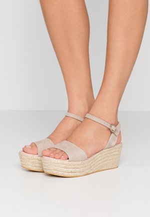 Sandales compensées en daim beige avec brides à la cheville portées pieds nus sur un fond blanc uni.