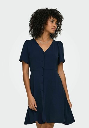 Kvinde med krøllet hår iført en marineblå kortærmet skjortekjole med knapper foran og sølvhalskæde, ser mod højre mod en ensfarvet baggrund.