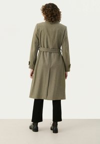 Oliivroheline villane trench coat, millel on vöökoht, tagumine vent ja nööbiga mansetid, koos mustade sametpükste ja mustade kingadega.