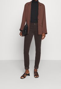Bruine oversized blazer, zwarte coltrui, donkerbruine slim-fit broek en zwarte sandalen met blokhak. Zwarte clutch in de hand.