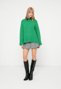 Zelený veľký pletený sveter s rebrovaným lemom, v kombinácii so sivou plisovanou sukňou a čiernymi čižmami nad kolená, na jednoduchom pozadí.