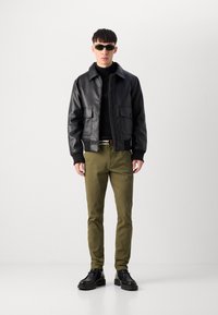 Jachetă bomber din piele neagră, cu manșete ribbate, pulover negru pe gât, pantaloni verzi măslinii și pantofi negri cu șireturi. Ochelari de soare supradimensionați verzi.