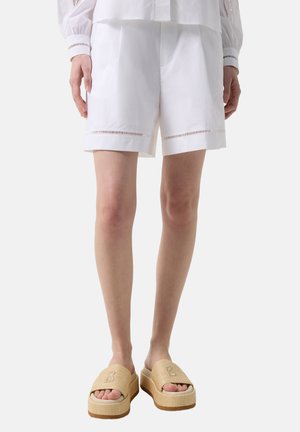 Frau trägt weiße, maßgeschneiderte Shorts mit dekorativen Ausschnitten am Saum und beige Plateausandalen auf neutralem Hintergrund.