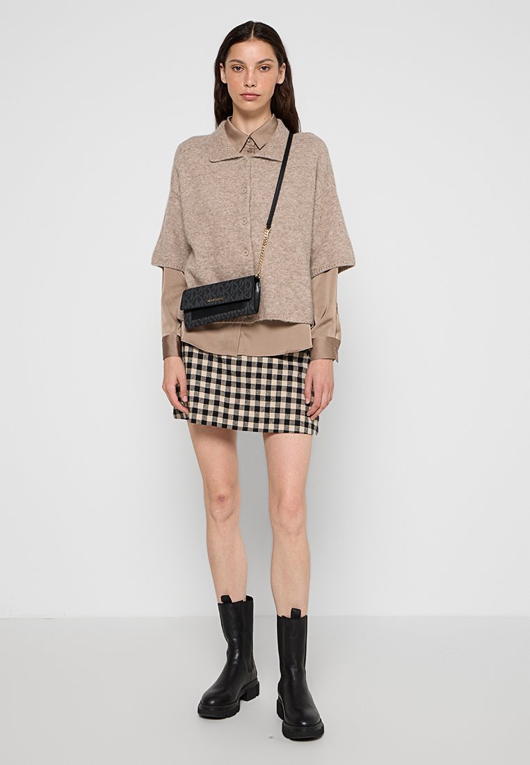 Beige wollen blend oversized top met knoopsluiting, gelaagd over een bruine blouse, gecombineerd met een zwart-wit geruit rokje en zwarte enkellaarsjes.