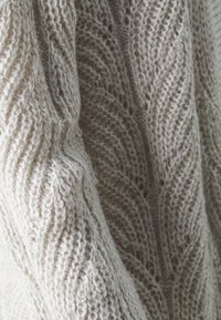 Beige gestrickter Pullover mit einer gerippten Textur, der ein Blättermuster und variierende Maschengrößen aufweist, was Weichheit und Wärme betont.