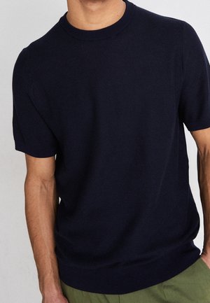 Man wearing a plain navy blue short-sleeve sweater with a crew neck and olive green pants, standing against a light background.