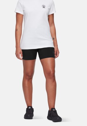 Persona con camiseta blanca de manga corta con un pequeño logo y pantalones cortos ajustados negros, de pie con zapatillas deportivas negras sobre fondo blanco.