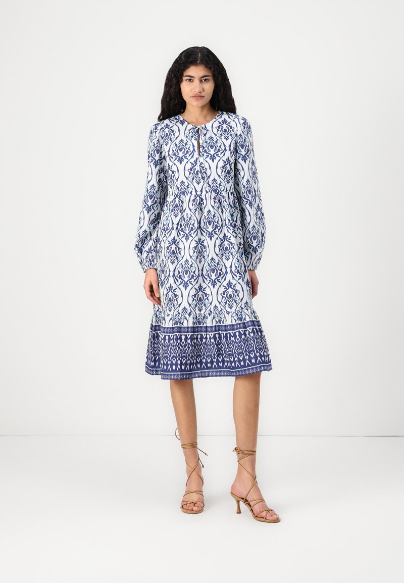 Rochie cu mâneci lungi, de lungime până la genunchi, având un model floral alb și albastru, guler rotund și margine evazată. Asortată cu sandale cu barete.