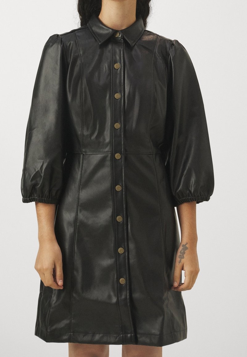 Femme portant une robe noire en similicuir avec des manches trois-quarts bouffantes et une rangée de boutons métalliques sur le devant.