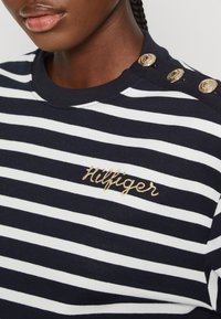 Sweatshirt rayé bleu marine et blanc avec broderie dorée "Hilfiger". Il présente quatre accents de boutons dorés sur l'épaule et un col côtelé.