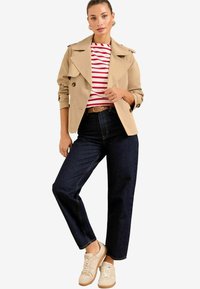 Beige Trenchcoat mit doppelreihigen Knöpfen, gestreiftem rot-weißem Hemd, dunkelblauen weit geschnittenen Jeans und beigen Sneakern mit strukturierten Akzenten.