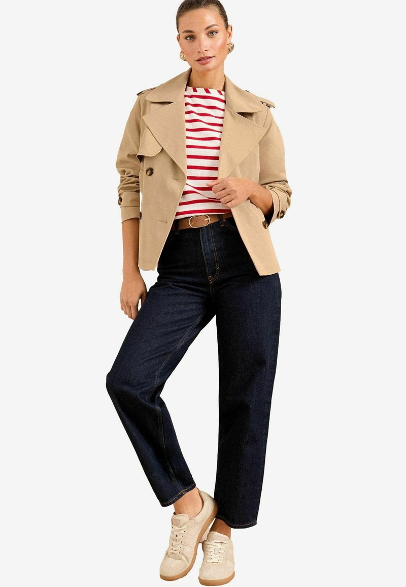 Beige Trenchcoat mit doppelreihigen Knöpfen, gestreiftem rot-weißem Hemd, dunkelblauen weit geschnittenen Jeans und beigen Sneakern mit strukturierten Akzenten.