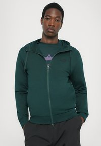 Zelená mikina na zips s kapucňou na šnúrku, vyrobená z mäkkého materiálu, s logom na hrudi a tesným strihom okolo pása a manžiet.