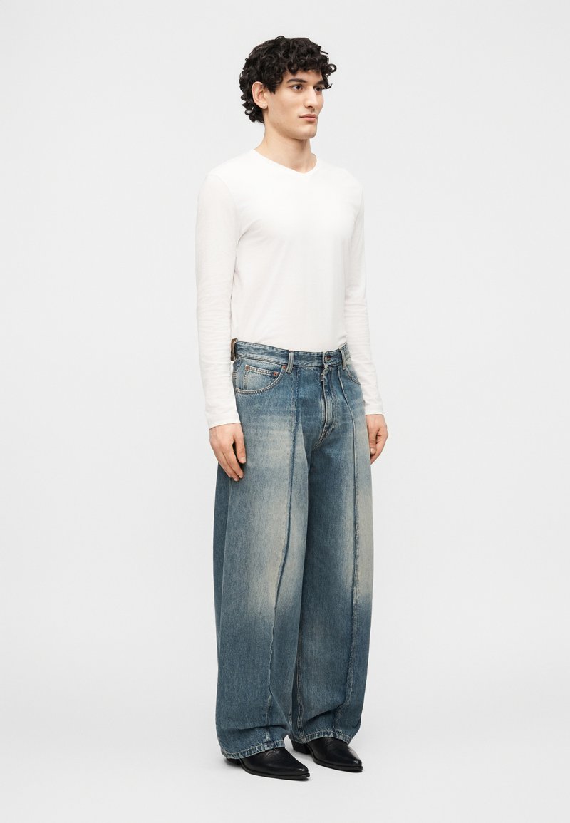 Man met krullend haar, gekleed in een wit shirt met lange mouwen, wijde verweerde blauwe jeans en zwarte puntige schoenen, staand tegen een effen achtergrond.