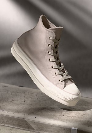 Sneaker alta in tela beige chiaro, con una punta in gomma bianca, lacci abbinati e una suola testurizzata con multiple righe orizzontali.