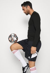 Schwarzes Langarmoberteil, schwarze Shorts mit weißen Akzenten, weiße Socken mit roten Streifen und schwarze Fußballschuhe. Spieler balanciert einen Fußball.
