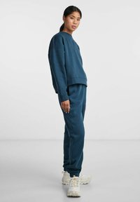 Dark teal sweatshirt and matching jogger pants made of textured fabric, featuring a relaxed fit, ribbed cuffs, and elastic waistband, paired with white sneakers.