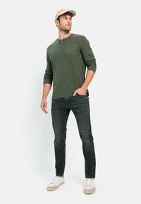 Olijfgroene lange mouwen henley-shirt met een zak, donkere spijkerbroek en witte sneakers. Het geheel wordt afgemaakt met een beige baseballpet.